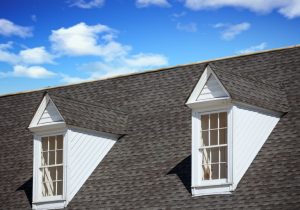 Home with grey asphalt shingles, two dormer windows, and a background with blue sky with cloud