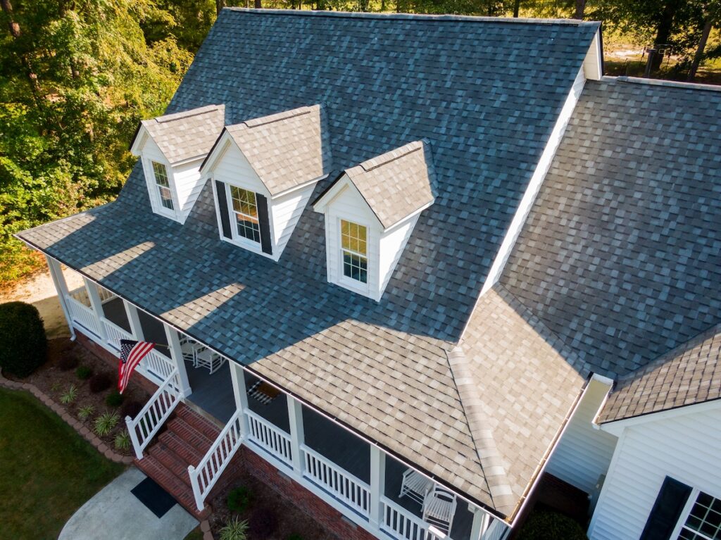 Aerial view of a grey-shingled, white house featuring three dormer windows and a large front porch with white railings—an inviting example of the types of roofing materials that enhance charm. Two rocking chairs, an American flag, and green trees complete the scene.