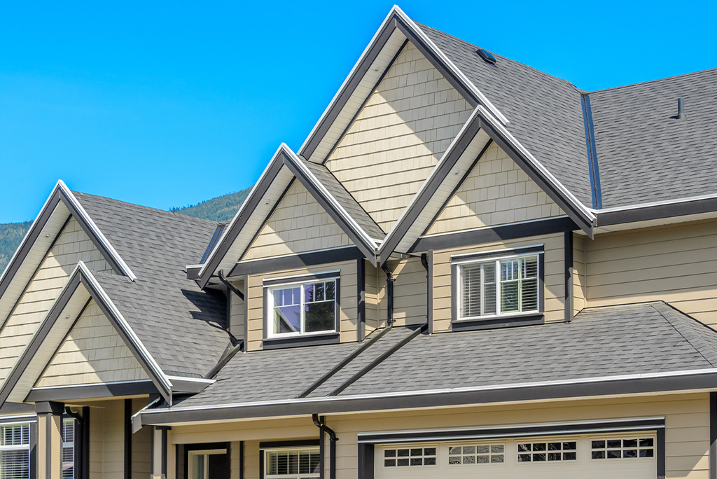 A close-up view of a modern house exterior with multiple gabled rooflines, showcasing various types of roofing materials, beige siding, and white-trimmed windows under a clear blue sky.