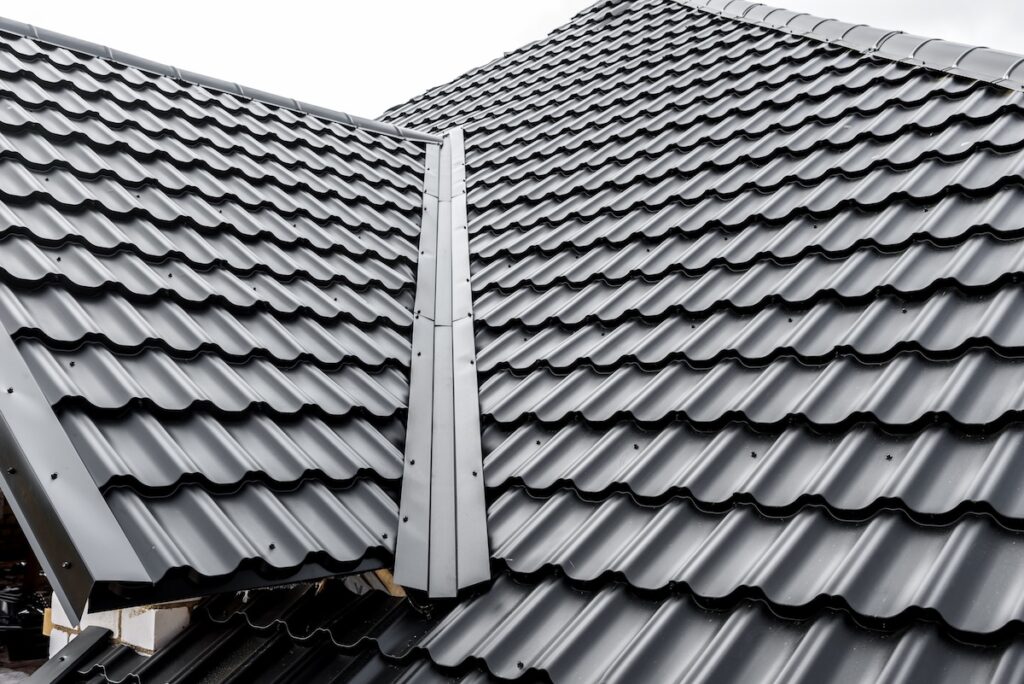 Close-up view of a house roof featuring dark, wavy metal roofing tiles—one of the top types of roofing materials—with a central metal ridge covering the roof joint, showcasing a modern and durable design against a cloudy sky.