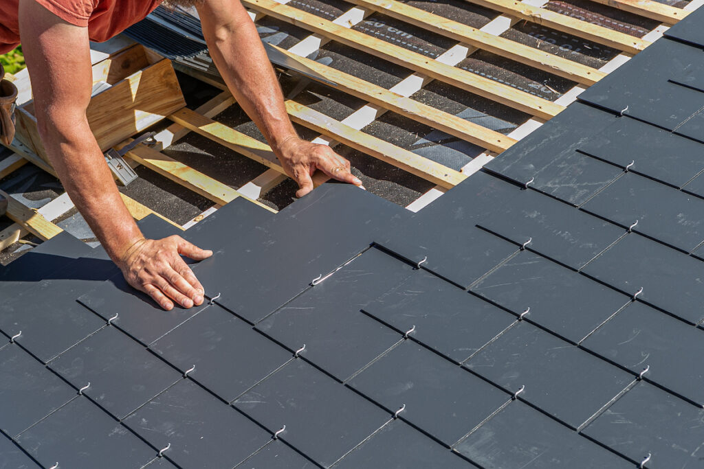 A person installs gray roof shingles—one of many types of roofing materials—on a wooden frame, using both hands to position the shingles in neat, overlapping rows. The image shows part of the roof under construction.