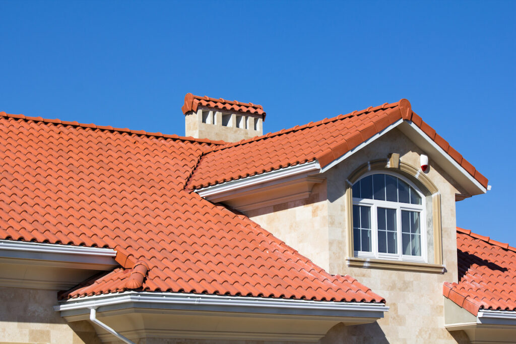 A close-up of a house with beige stone walls and a red tiled roof—one of the classic types of roofing materials—featuring a white arched window and clear blue sky in the background.