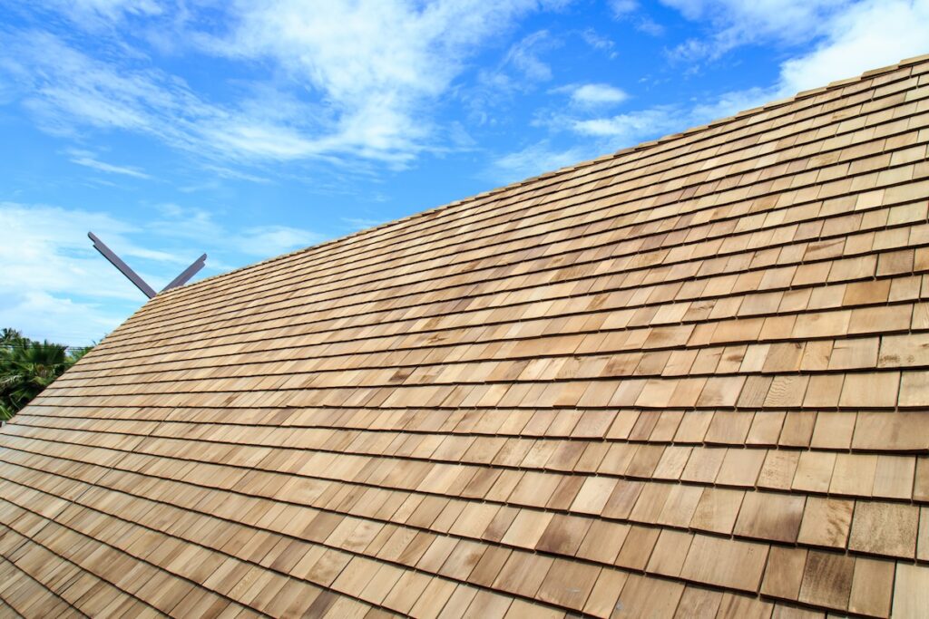 Wooden shingles, one of the classic types of roofing materials, cover a steeply sloped roof under a bright blue sky with scattered clouds. The well-constructed roof looks new, with trees barely visible in the corner.