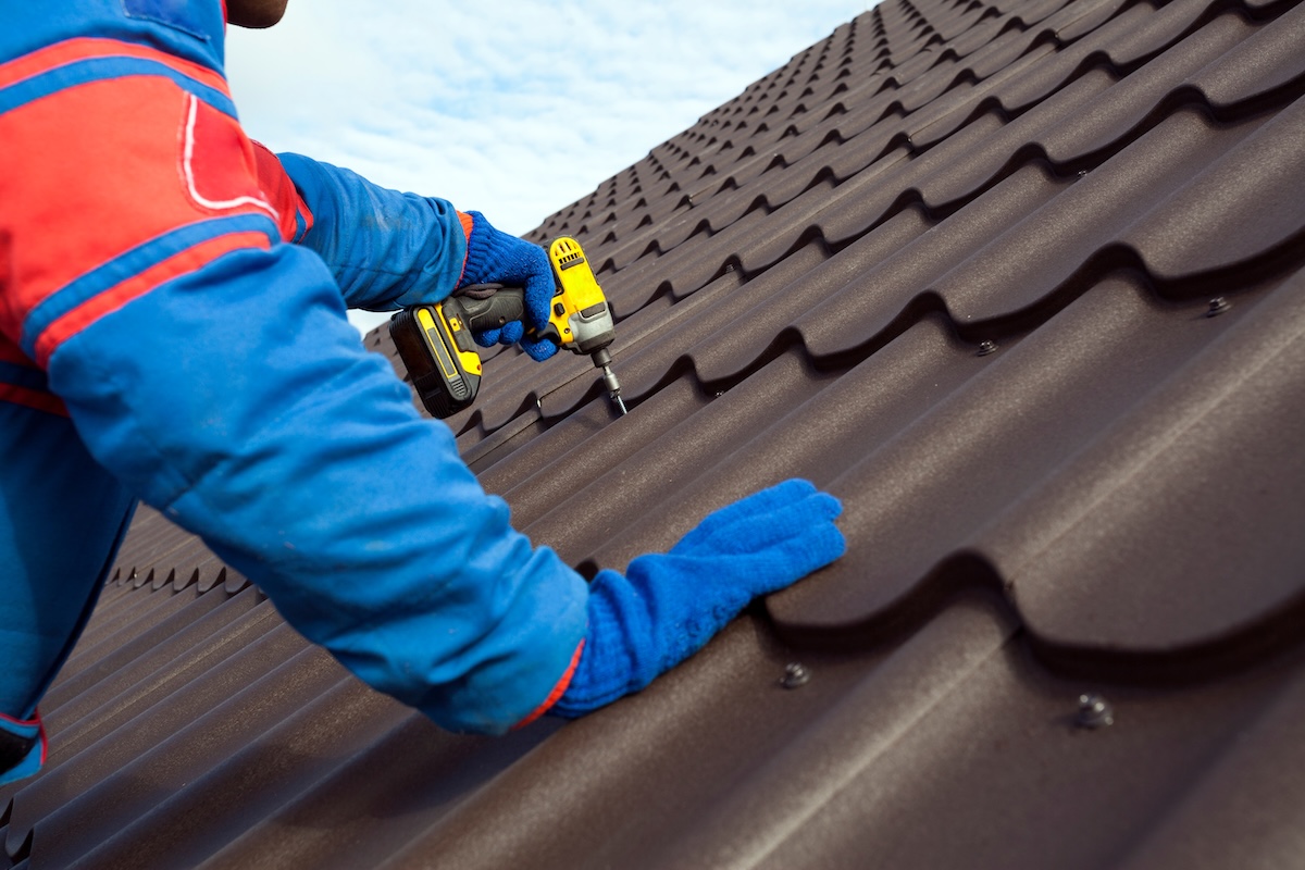 A worker in blue gloves and a protective suit uses a yellow power drill to secure dark sheets, showcasing one of the popular types of metal roofing