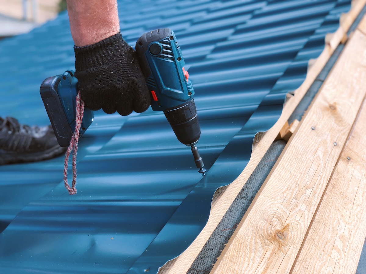 A gloved hand uses a power drill to fasten a screw into blue corrugated metal roofing sheets