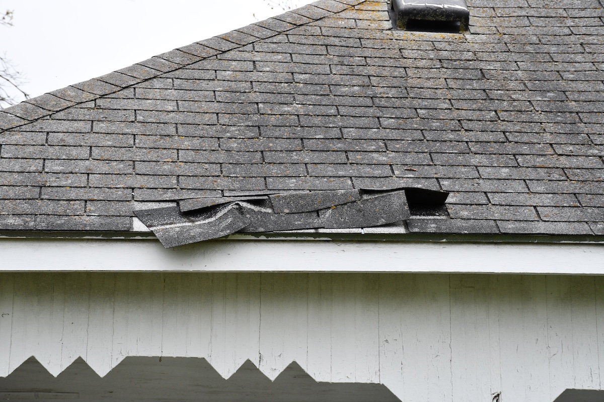 A close-up of a house roof with several loose, curled asphalt shingles near the edge,