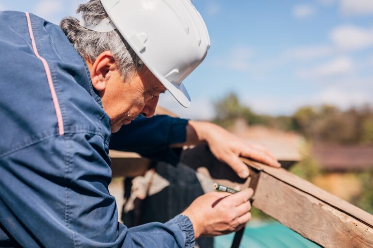 A construction worker in a white hard hat and blue jacket measures a wooden beam outdoors