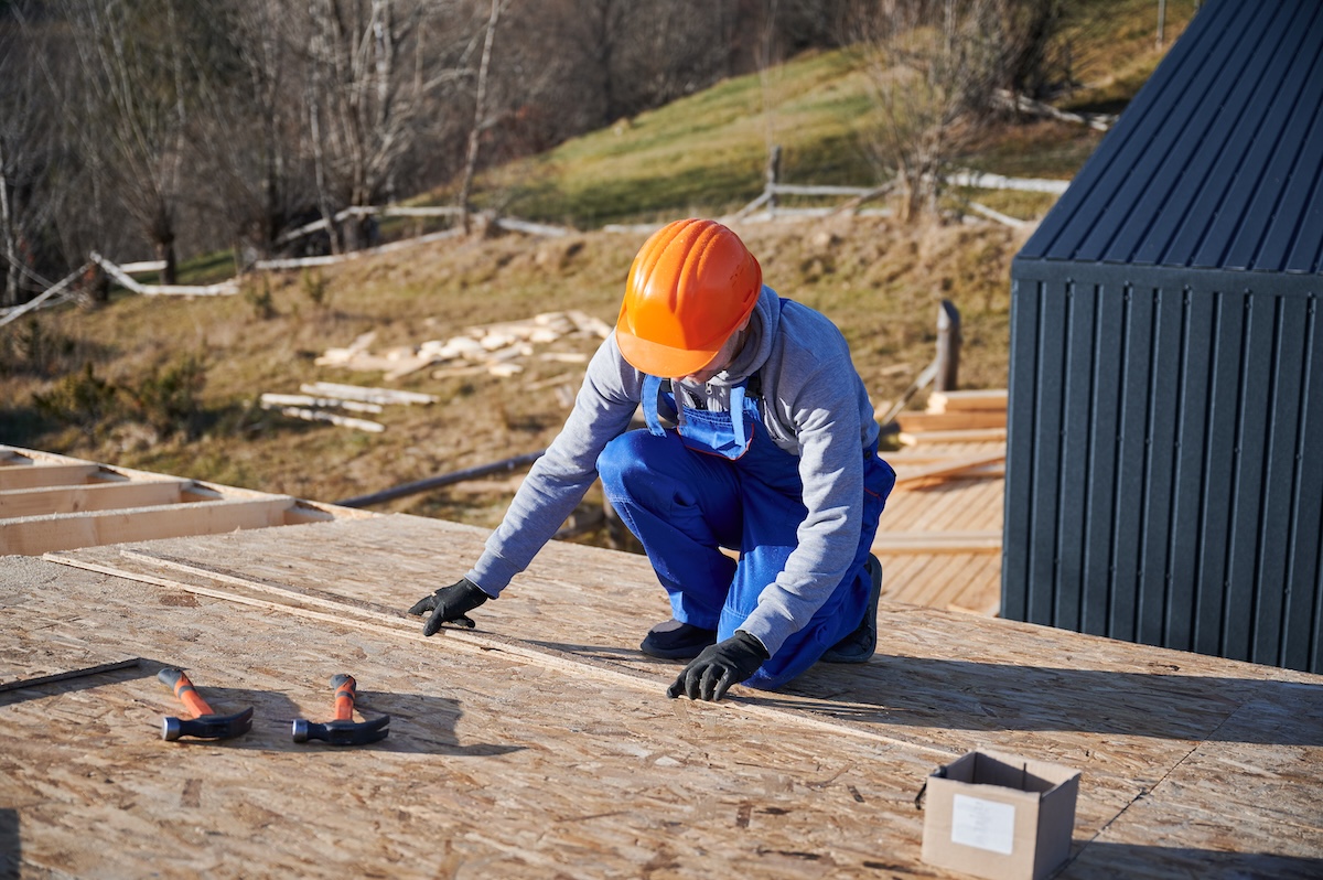 A construction worker kneels on a wooden roof, measuring
