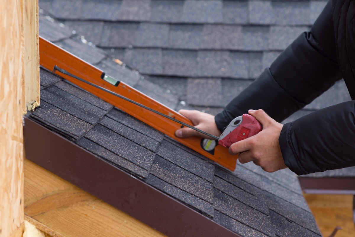 A person uses a spirit level and tape measure to check the angle of a shingled roof.
