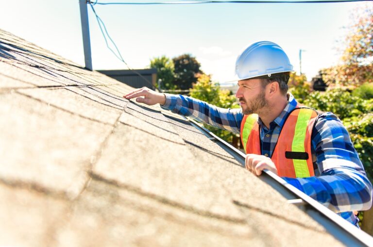 A man wearing a hard hat and safety vest inspects the shingles on a house roof