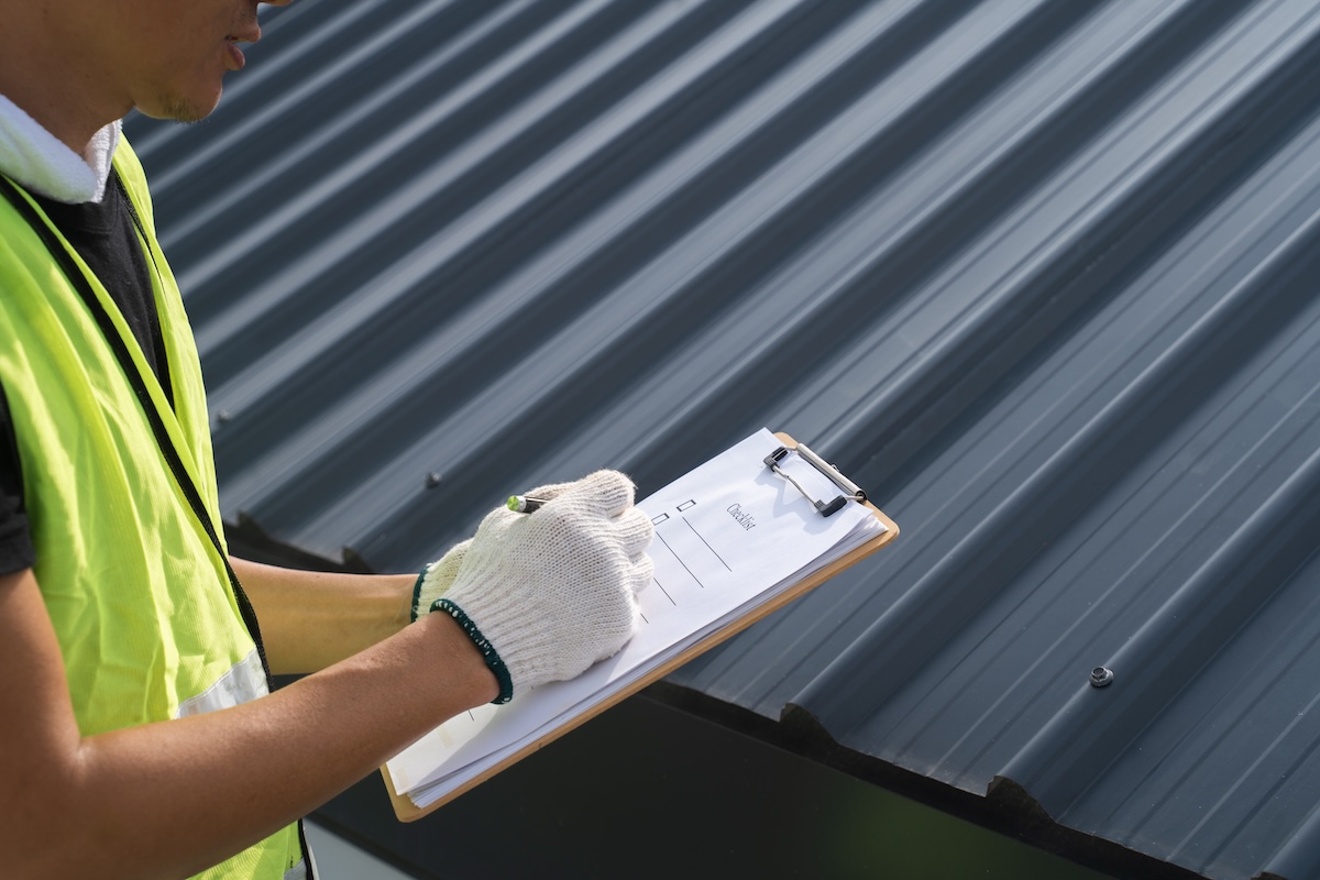 A person in a yellow safety vest and white gloves holds a clipboard and pen, inspecting a dark corrugated metal roof up close